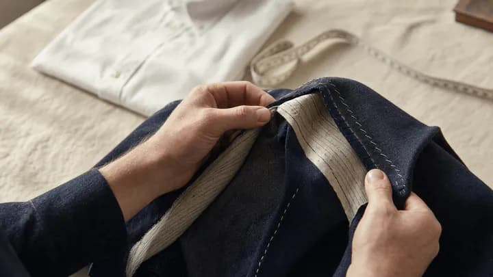 Close-up of hands inspecting the inside seams and lining of a navy wool jacket on a linen-covered table in soft daylight.