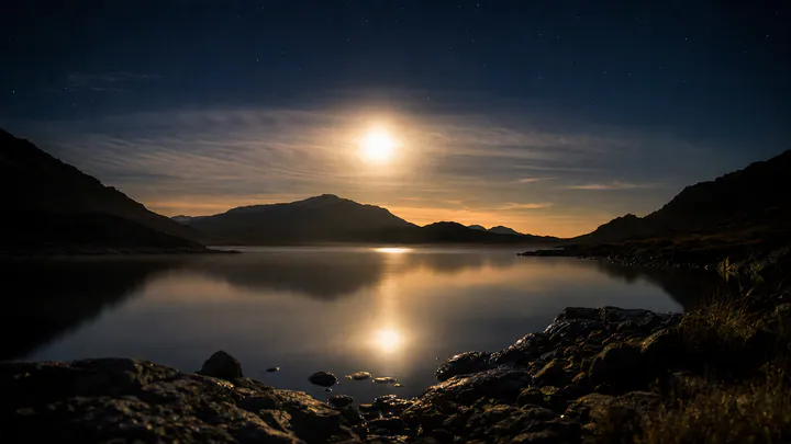 A bright full moon rising over dark hills, haloed by thin cloud and reflected in still water under an indigo sky.
